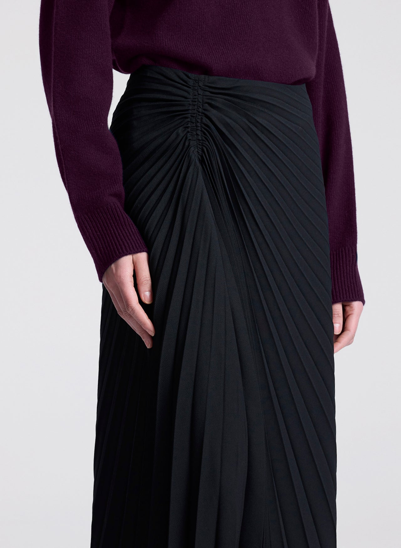 Person wearing a dark purple sweater and black pleated skirt on a light gray background